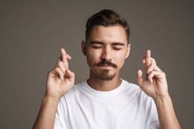 Şans dilemek için parmaklarını çaprazlayarak poz veren gergin, tıraşsız adamın görüntüsü gri arka planda izole edilmiş.