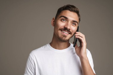 Gri arka planda izole edilmiş, gülümseyen ve cep telefonuyla konuşan neşeli, tıraşsız adamın görüntüsü.