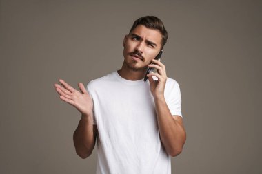 Gri arka planda izole bir şekilde cep telefonuyla konuşurken kafası karışmış bir adamın görüntüsü