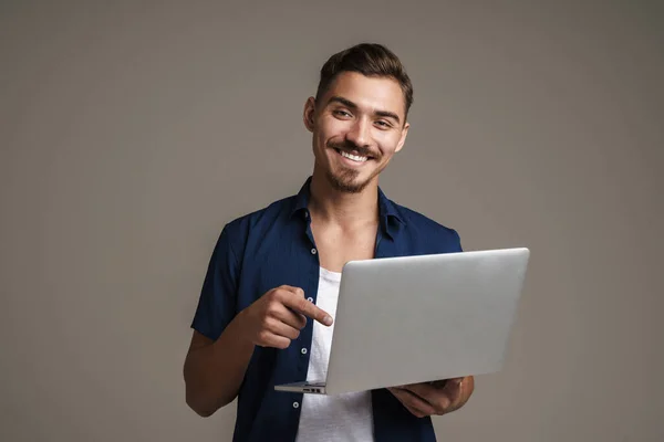 Gri Arkaplanda Dizüstü Bilgisayarı Tutan Işaret Eden Mutlu Yakışıklı Adam — Stok fotoğraf