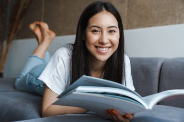 Evde oturup dergi okuyan neşeli, genç bir kadının görüntüsü.