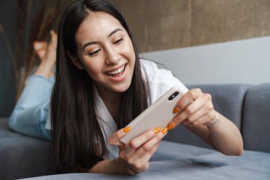 Evde, kanepede uzanmış, cep telefonuyla oyun oynayan mutlu genç bir kadın.