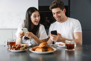 Mutfakta lezzetli bir kahvaltı yaparken cep telefonu kullanan mutlu genç bir çiftin portresi.
