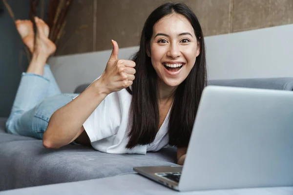 Happy young woman on a video call through laptop computer while laying on a couch at home, thumbs up