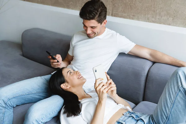 一对快乐的夫妇一边在智能手机里享受媒体的内容 一边在家里的沙发上放松一下 — 图库照片
