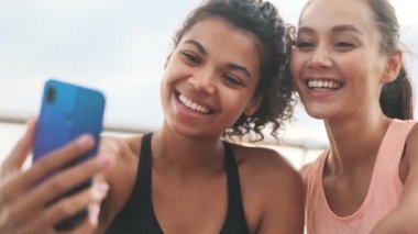 Sahilde cep telefonuyla fotoğraf çeken neşeli genç spor kadınları.