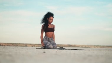 Ciddi bir Afro-Amerikalı kadın spor kıyafetleri giyiyor. Kollarını esnetiyor. Sahildeki yoga minderinin üzerinde oturuyor.