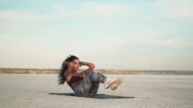 Spor kıyafetleri giyen çekici, Afro-Amerikalı bir kadın plajdaki yoga minderinde antrenman yapıyor.