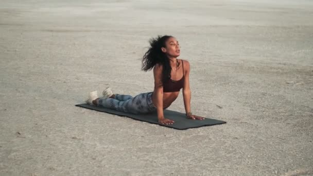 一位身穿运动服的专注的年轻非洲裔美国女性正在海滩外的瑜伽垫上做伸展运动 — 图库视频影像