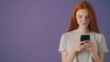 Memnun genç bir kadın, menekşe arka planında izole bir şekilde duran akıllı telefonunu kullanıyor.