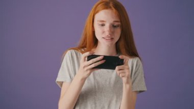 Gülümseyen genç bir kadın akıllı telefonuyla oyun oynuyor menekşe arka planda tek başına duruyor.