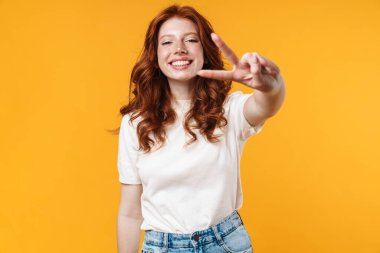 Kızıl, neşeli, göz kırpan ve işaret eden kızın görüntüsü sarı arka planda izole edilmiş.