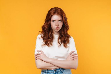 Kızıl, hoşnutsuz, kolları çaprazlanmış bir kız görüntüsü sarı arka planda izole edilmiş.