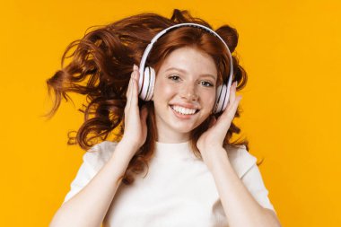 Kızıl saçlı heyecanlı kızın sarı arka planda izole edilmiş kulaklık kullanması.