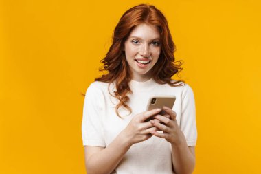 Sarı arka planda izole edilmiş, gülümseyen ve cep telefonu kullanan kızıl mutlu bir kız resmi