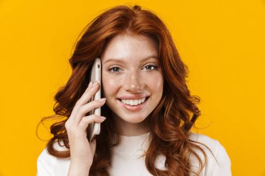 Sarı arka planda izole edilmiş, gülümseyen ve cep telefonuyla konuşan kızıl mutlu kız görüntüsü