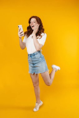 Kızıl saçlı heyecanlı kız görüntüsü şaşırırken sarı arkaplanda izole edilmiş cep telefonu kullanıyor