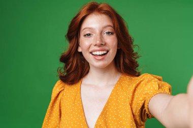 Yeşil arka planda izole edilmiş gülümseyen ve selfie çeken kızıl saçlı bir kız resmi