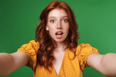 Kızıl saçlı şaşırmış bir kızın yeşil arka planda izole edilmiş selfie fotoğrafını çekmesi.