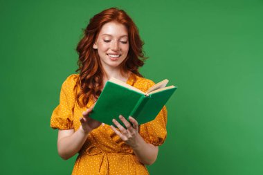 Yeşil arka planda izole edilmiş gülümseyen ve kitap okuyan kızıl saçlı neşeli kız görüntüsü