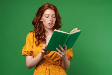 Kızıl saçlı kızın yeşil arka planda izole edilmiş kitap okumasına şaşırmış görünümü