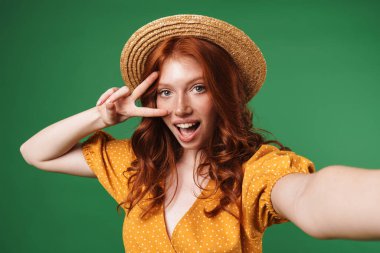 Saman şapkalı neşeli kızıl bir kız resmi barış işareti yapıyor ve yeşil arka planda izole edilmiş selfie çekiyor.
