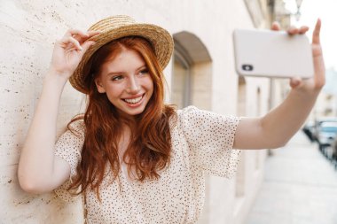 Hasır şapkalı, gülümseyen kızıl bir kızın cep telefonuyla fotoğraf çekmesi. Şehir sokaklarında yürürken.