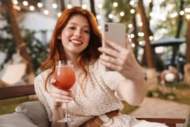 Gülümseyen kızıl kızın cep telefonuyla selfie çekmesi ve yaz kafesinde oturup kokteyl içmesi.