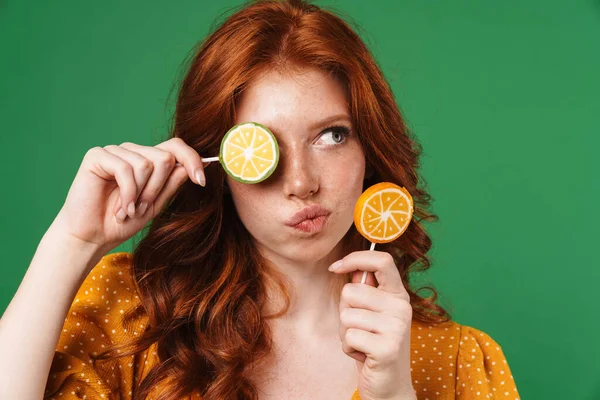 Image of redhead thinking girl posing with lollipops isolated over green background