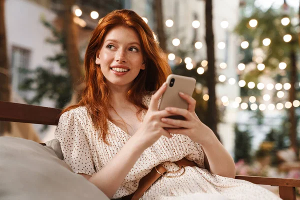Зображення Задоволеної Імбирної Дівчини Яка Посміхається Використовує Мобільний Телефон Сидячи — стокове фото