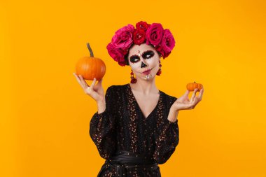 Cadılar Bayramı makyajlı hoşnutsuz genç kız sarı arka planda izole edilmiş balkabaklarıyla poz veriyor.