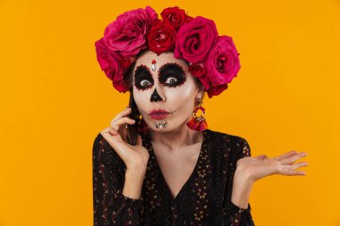 Cadılar Bayramı makyajı ve çiçek çelengi giymiş kafası karışmış bir kız sarı duvarın üstünde cep telefonuyla konuşuyor.