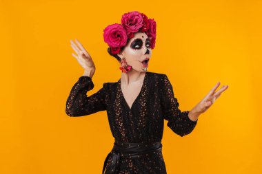 Cadılar bayramı makyajı ve çiçek çelengi giymiş şaşırmış kız sarı duvarın üstünde dans ediyor.