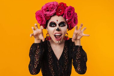 Cadılar bayramı makyajı ve çiçek çelengi giymiş ürkütücü kız sarı duvarda izole edilmiş kameraya poz veriyor.