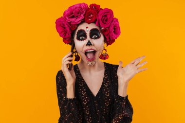 Cadılar bayramı makyajı ve çiçek çelengi giymiş heyecanlı kız sarı duvarda izole bir şekilde cep telefonundan konuşuyor.