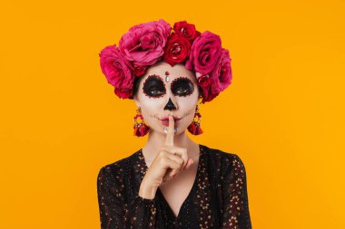 Cadılar bayramı makyajı ve çiçek çelengi içinde güzel bir kız sarı duvarın üzerinde sessiz bir jest yapıyor.