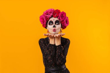 Cadılar bayramı makyajı ve çiçek çelengi giymiş mutlu bir kız. Sarı duvarın üzerinde yalıtılmış bir şekilde öpüşüyor.