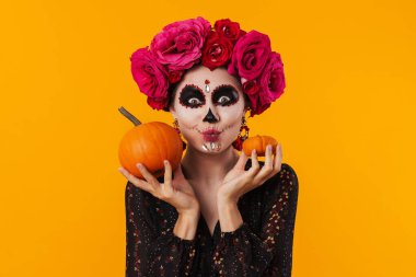 Cadılar Bayramı makyajlı neşeli genç kız sarı arka planda izole edilmiş balkabaklarıyla poz veriyor.