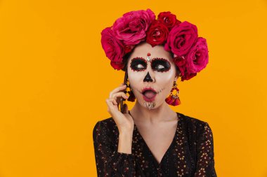 Cadılar Bayramı makyajı ve çiçek çelengi giymiş kafası karışmış bir kız sarı duvarın üstünde cep telefonuyla konuşuyor.