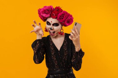 Cadılar Bayramı makyajlı korkunç kız ve sarı duvarda izole edilmiş cep telefonu kullanan çiçek çelengi.