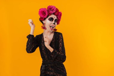 Cadılar bayramı makyajı ve çiçek çelengi giymiş neşeli kız sarı duvarın üstünde dans ediyor.