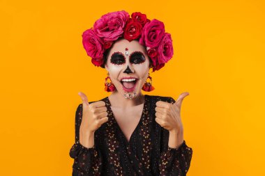 Cadılar Bayramı makyajı ve çiçek çelengi içindeki heyecanlı kız sarı duvarın üzerinde parmaklarını gösteriyor.
