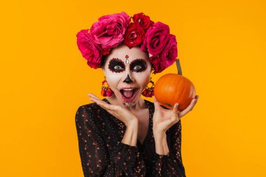 Cadılar Bayramı makyajlı heyecanlı genç kız sarı arka planda izole edilmiş balkabağıyla poz veriyor.