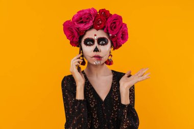 Cadılar Bayramı makyajı ve çiçek çelengi giymiş kafası karışmış bir kız sarı duvarın üstünde cep telefonuyla konuşuyor.