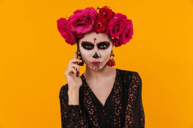 Cadılar Bayramı makyajı ve çiçek çelengi giymiş hoşnutsuz bir kız sarı duvarın üstünde cep telefonuyla konuşuyor.