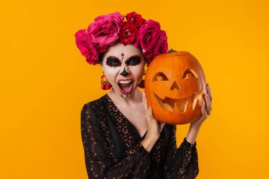 Cadılar Bayramı makyajlı korkunç genç kız sarı arka planda izole edilmiş balkabağıyla poz veriyor.