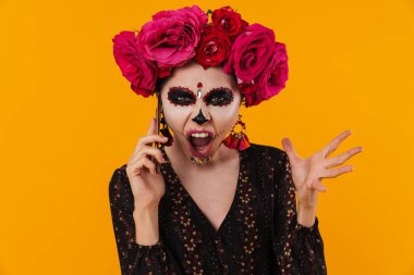 Cadılar bayramı makyajı ve çiçek çelengi giymiş kızgın kız sarı duvarda izole bir şekilde cep telefonundan konuşuyor.