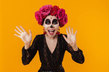 Cadılar bayramı makyajı ve çiçek çelengi giymiş heyecanlı kız sarı duvarda izole edilmiş kameraya poz veriyor.