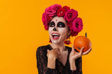 Cadılar Bayramı makyajlı, sarı arka planda balkabağıyla poz veren gülen genç kız.