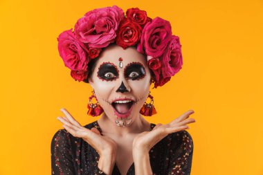 Cadılar bayramı makyajı ve çiçek çelengi giymiş mutlu bir kız sarı duvarda izole edilmiş kameraya poz veriyor.